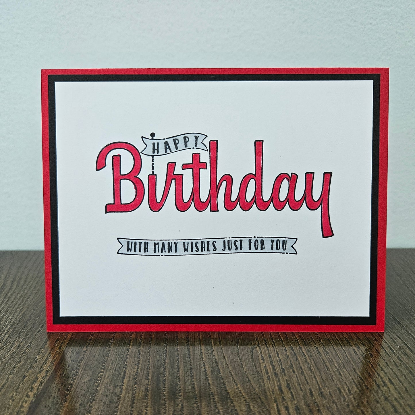 A square handmade greeting card with a red border, black inner border, and red script text reading 'Happy Birthday' with a smaller black banner below, displayed on a wooden surface against a light wall.