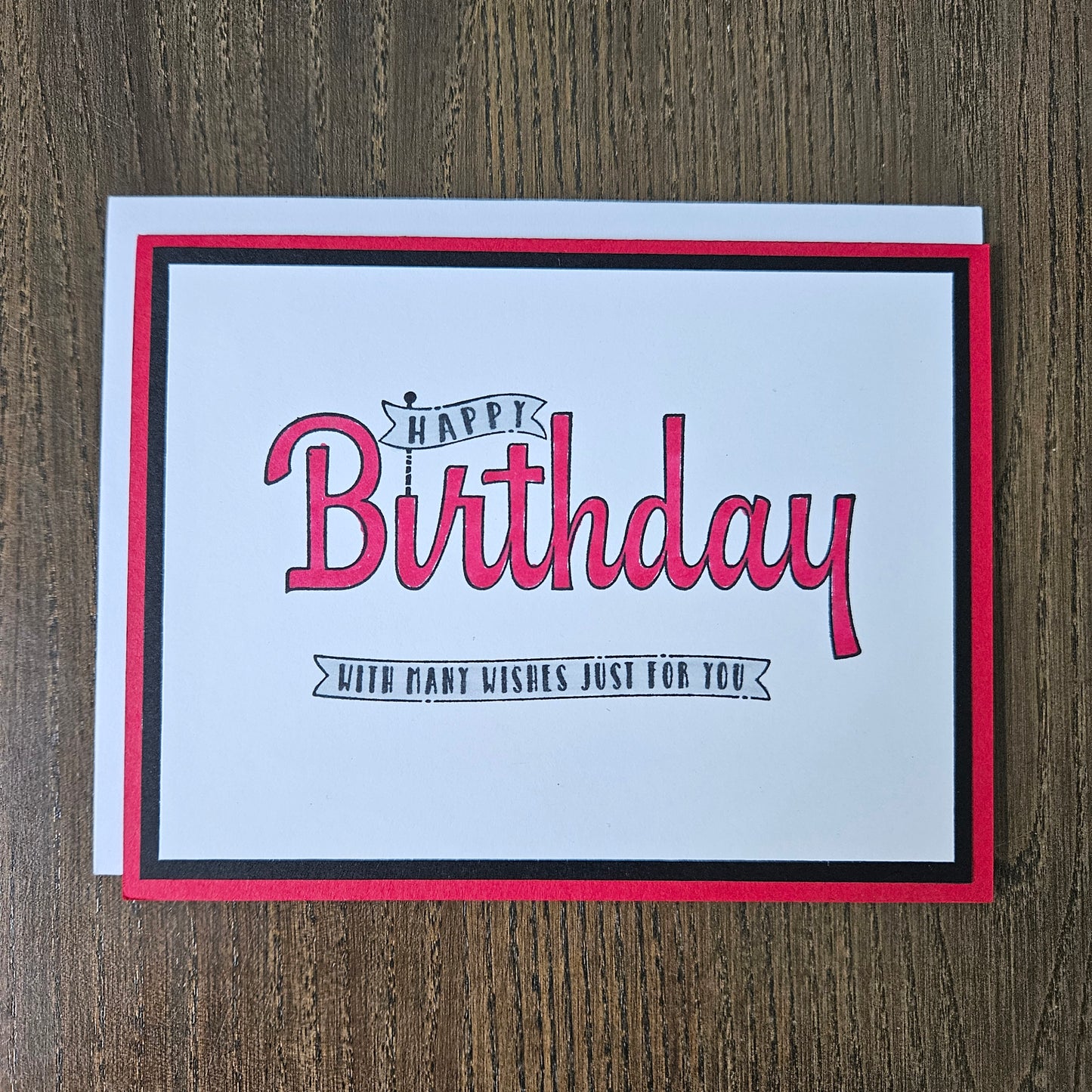 A square birthday card with a white center, red and black border, and pink script text reading 'Happy Birthday' with smaller text 'With many kisses just for you,' photographed on a brown wood surface.