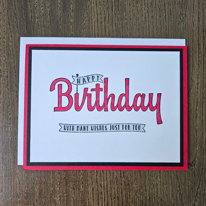 A square birthday card with a white center, red and black border, and pink script text reading 'Happy Birthday' with smaller text 'With many kisses just for you,' photographed on a brown wood surface.