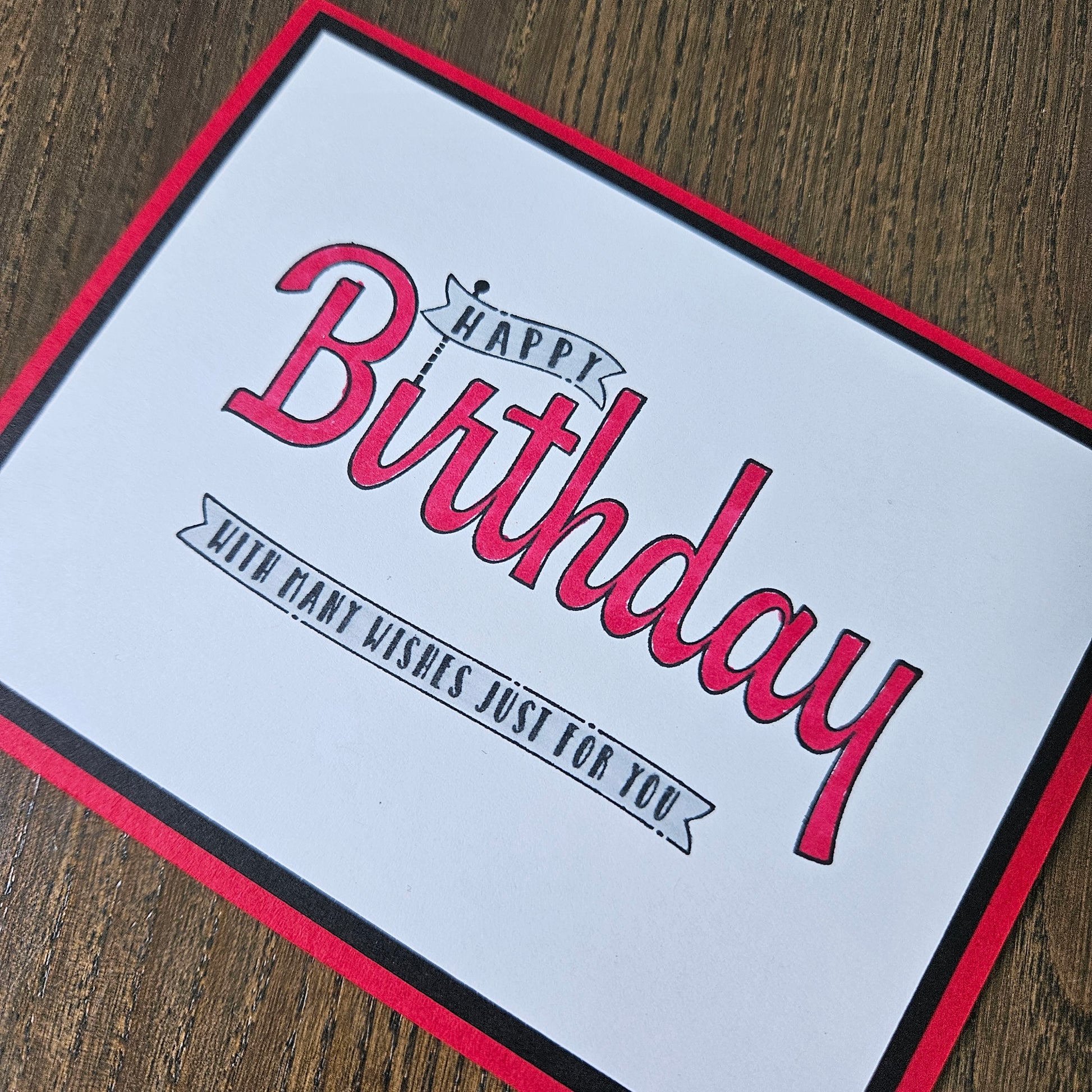 A square birthday card with a white center, red border, and black trim, featuring the words 'Happy Birthday' in red script and a smaller banner reading 'With my wishes just for you.' The card is photographed on a wooden surface.