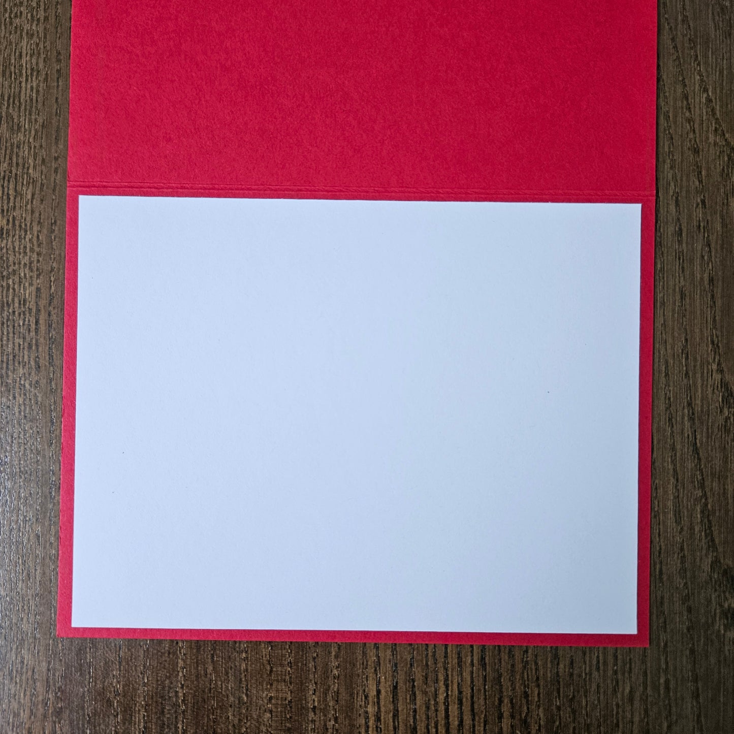 A red greeting card or notebook cover partially open, showing a blank white insert page on a dark wood background.