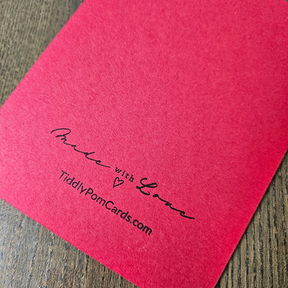 A close-up of a bright pink textured card or paper sheet placed on a wooden surface, with black printed text reading 'Made with Love' and 'TidilyPomCards.com'.