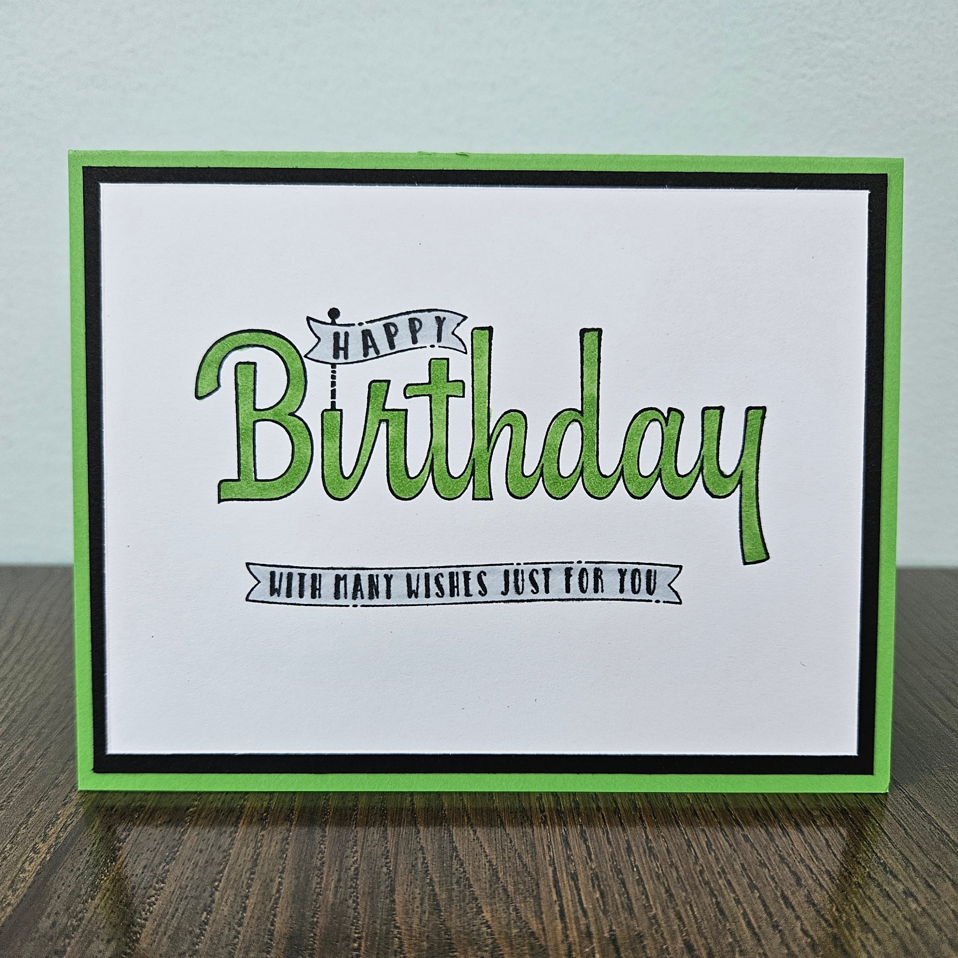 A handmade square greeting card with a white center, green and black borders, and the words “Happy Birthday with many wishes just for you” in green and black lettering, displayed on a dark wood surface against a light wall.