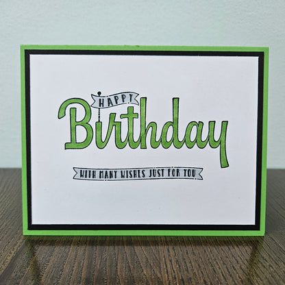 A handmade square greeting card with a white center, green and black borders, and the words “Happy Birthday with many wishes just for you” in green and black lettering, displayed on a dark wood surface against a light wall.