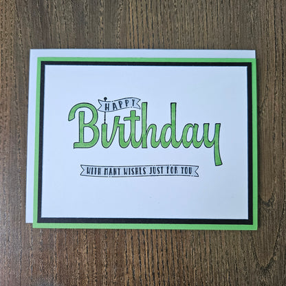 A square birthday greeting card with a white center, green and black border, and the words “Happy Birthday” in green script, placed on a brown wood surface.