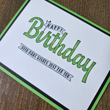 A square greeting card with a white center, green border, and green-and-black script text reading 'Happy Birthday' and 'Wish that wishes just for you,' photographed on a brown wood surface.