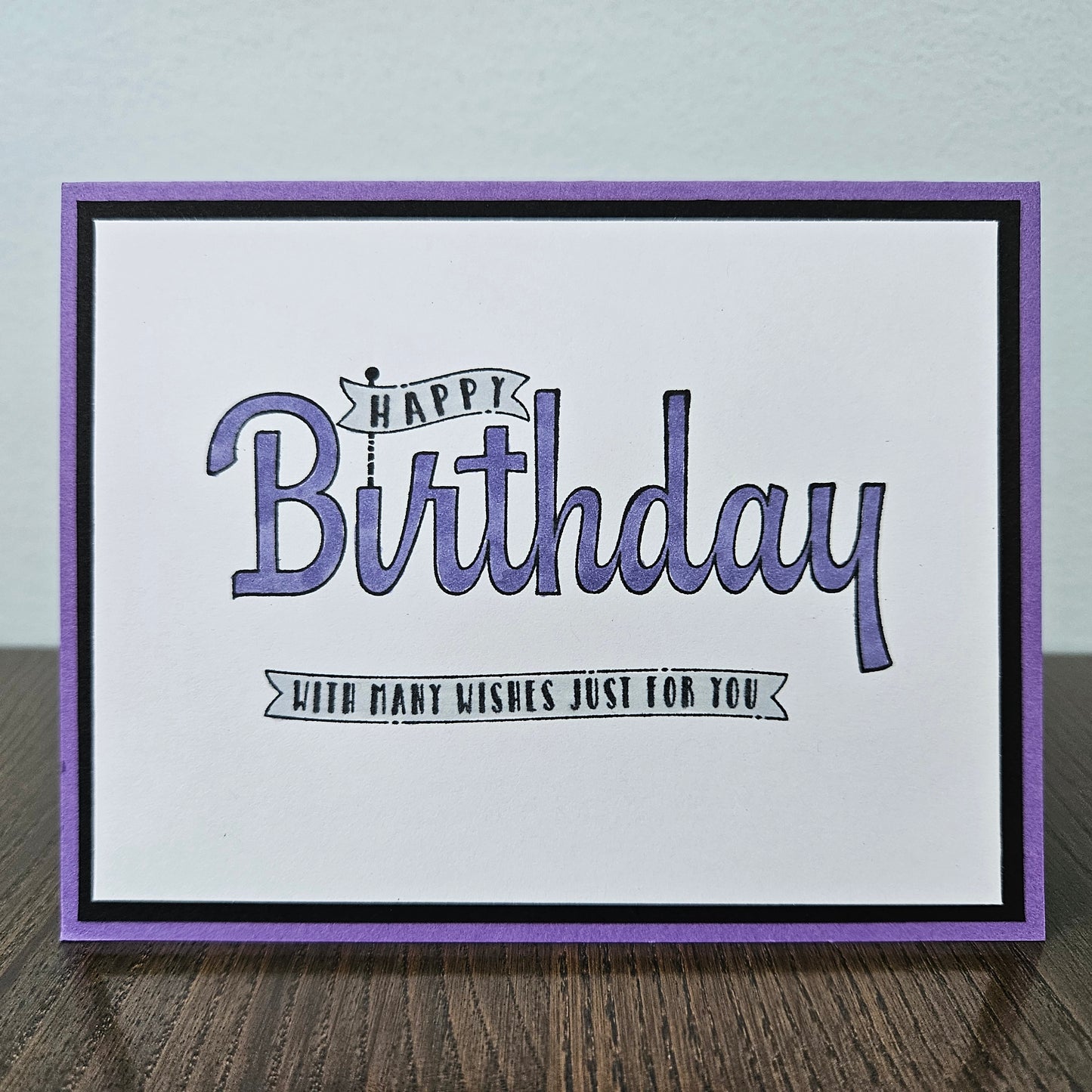 A handmade square greeting card with a purple border, black inner border, and a white center featuring the text 'Happy Birthday with many wishes just for you' in purple and black script, displayed on a wooden surface.