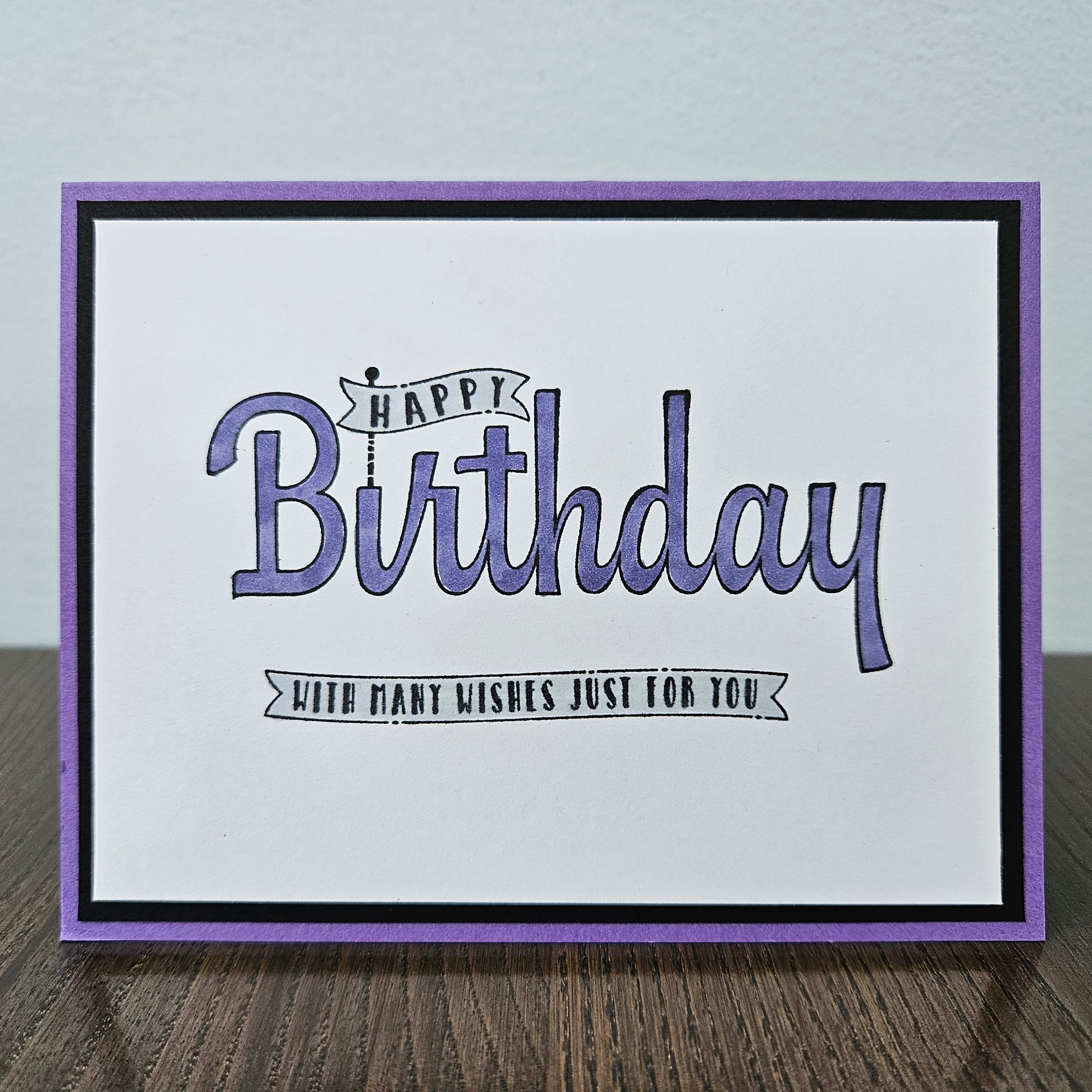 A handmade square greeting card with a purple border, black inner border, and a white center featuring the text 'Happy Birthday with many wishes just for you' in purple and black script, displayed on a wooden surface.