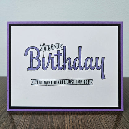 A handmade square greeting card with a purple border, black inner border, and a white center featuring the text 'Happy Birthday with many wishes just for you' in purple and black script, displayed on a wooden surface.