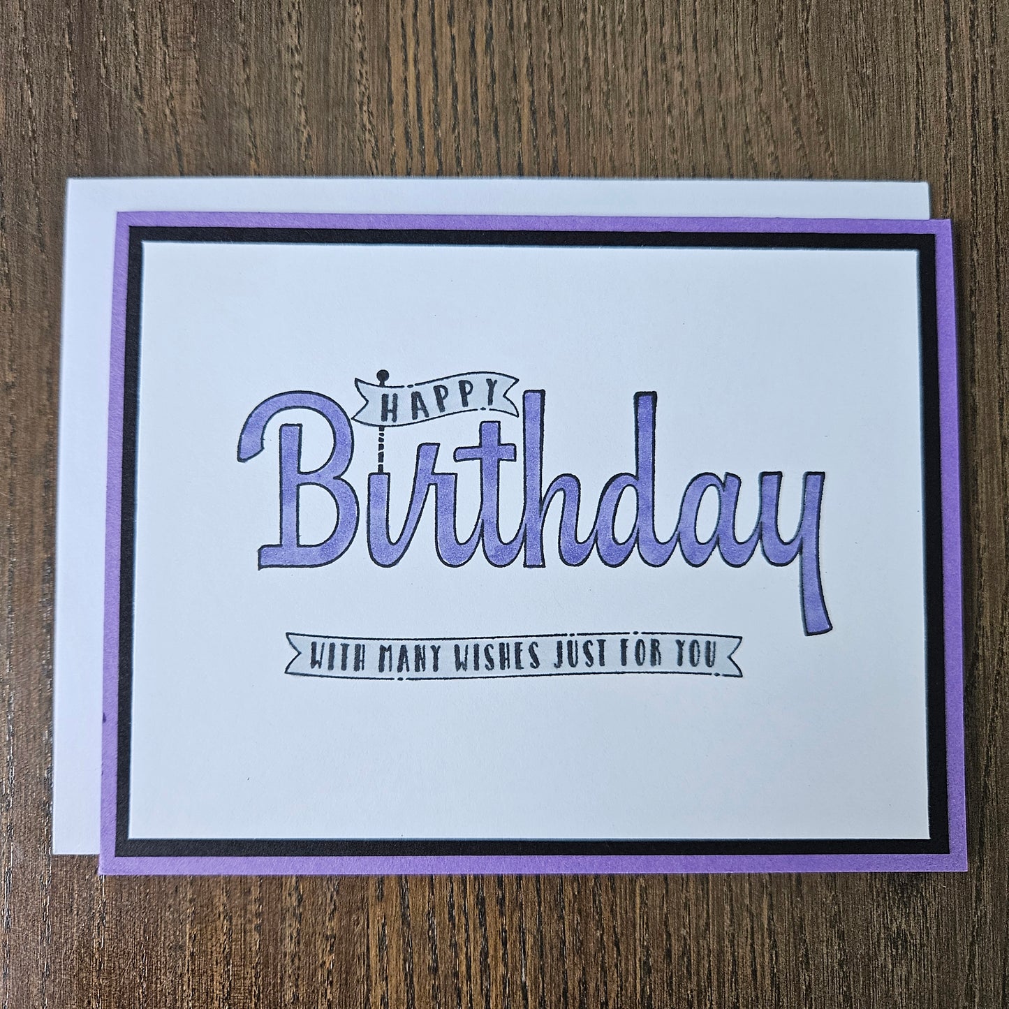 A square birthday card with a white center, purple border, and black inner frame, placed on a brown wood surface. The card reads “Happy Birthday” in large purple script with smaller text that says “JUDY’S FIRST WISHES JUST FOR YOU.