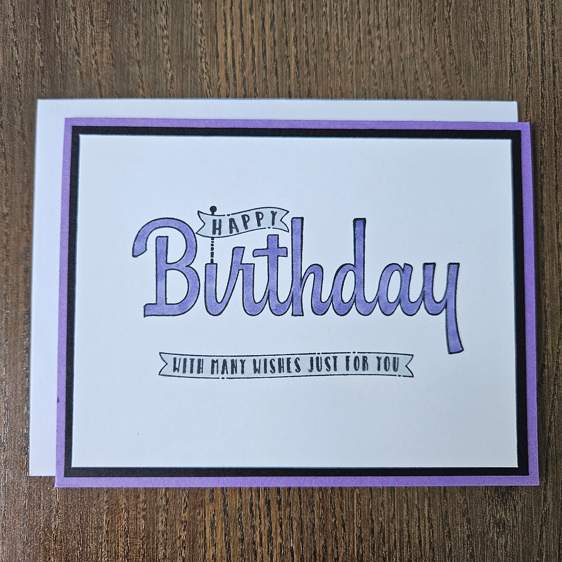 A square birthday card with a white center, purple border, and black inner frame, placed on a brown wood surface. The card reads “Happy Birthday” in large purple script with smaller text that says “JUDY’S FIRST WISHES JUST FOR YOU.