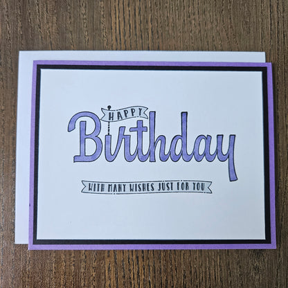 A square birthday card with a white center, purple border, and black inner frame, placed on a brown wood surface. The card reads “Happy Birthday” in large purple script with smaller text that says “JUDY’S FIRST WISHES JUST FOR YOU.