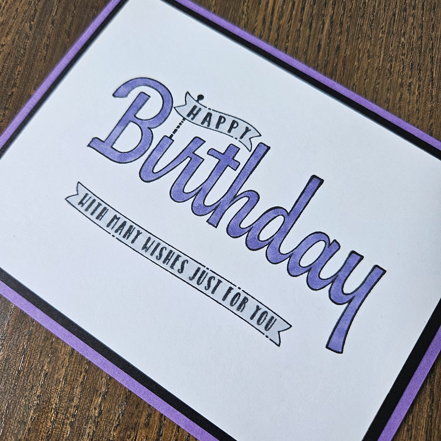 A square birthday card with a white center, purple border, and purple text reading 'Happy Birthday' and 'With warm wishes just for you,' photographed on a wooden surface.