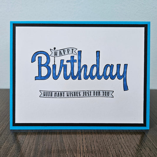 A square handmade greeting card with a blue border, black inner border, and a white center. The card features the text 'HAPPY Birthday' in blue script with a small banner detail, and 'WITH MANY WISHES JUST FOR YOU' in black script below, displayed on a dark wood surface against a light blue wall.