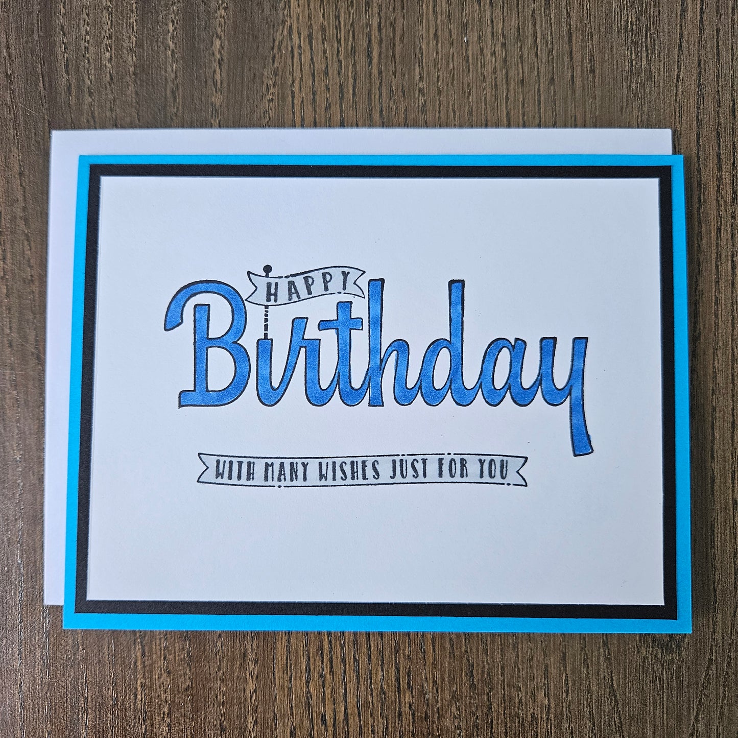 A square birthday greeting card with a white center, blue and black border, and blue script text reading 'Happy Birthday' with smaller text 'With many wishes just for you,' photographed on a wood surface.