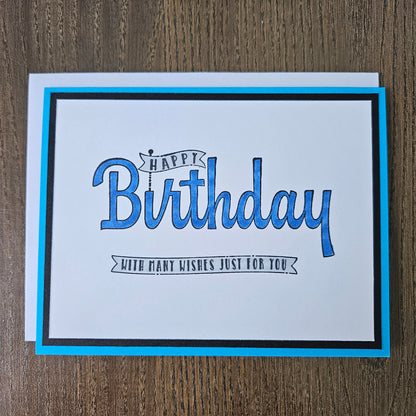 A square birthday greeting card with a white center, blue and black border, and blue script text reading 'Happy Birthday' with smaller text 'With many wishes just for you,' photographed on a wood surface.