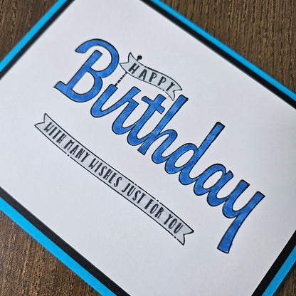 A close-up of a square birthday card with a white face, blue border, and blue-and-black script text reading 'Happy Birthday with many wishes just for you,' placed on a wooden surface.