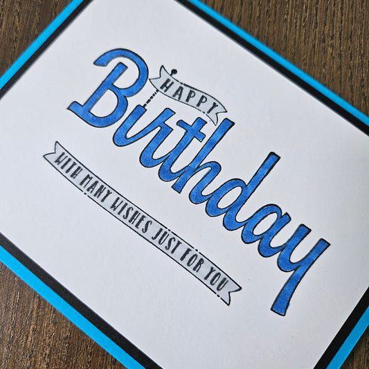 A close-up of a square birthday card with a white face, blue border, and blue-and-black script text reading 'Happy Birthday with many wishes just for you,' placed on a wooden surface.