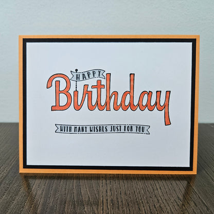 A square greeting card with a white center, orange and black script text reading 'Happy Birthday' and 'With many wishes just for you,' framed by a black inner border and an orange outer border, standing on a dark wood surface against a light gray wall.