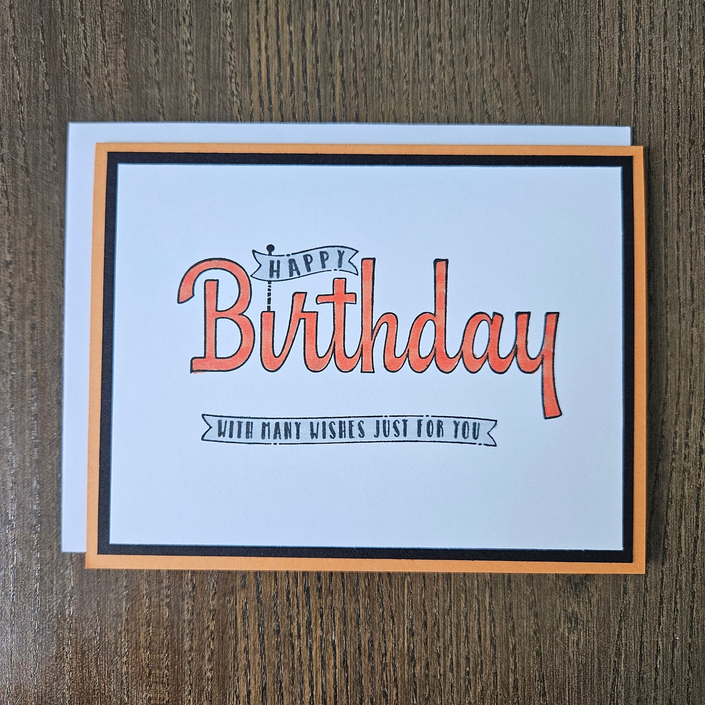 A square birthday card with a white center, orange and black border, and orange script text reading 'Happy Birthday' with smaller text 'With many wishes just for you,' photographed on a brown wood surface.