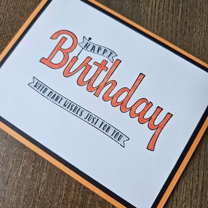 A square birthday card with a white background, featuring the words 'Happy Birthday' in orange script and 'With many wishes just for you' on a ribbon banner. The card has a black inner border and an orange outer border, and is photographed at an angle on a dark wood surface.