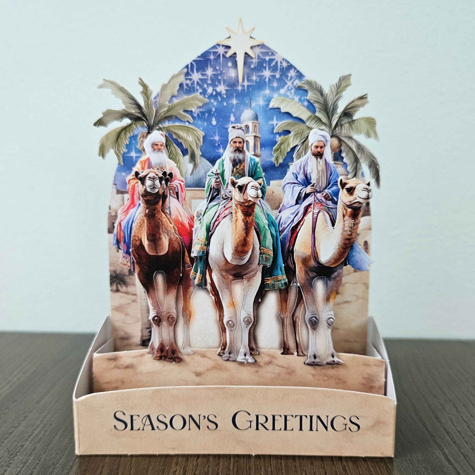 A handmade 3D pop-up cardboard Christmas greeting card displaying the Three Wise Men on camels under a starry blue sky, set in a display stand with 'SEASON'S GREETINGS' printed on the front, resting on a dark wooden surface.