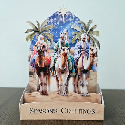 A handmade 3D pop-up cardboard Christmas greeting card displaying the Three Wise Men on camels under a starry blue sky, set in a display stand with 'SEASON'S GREETINGS' printed on the front, resting on a dark wooden surface.
