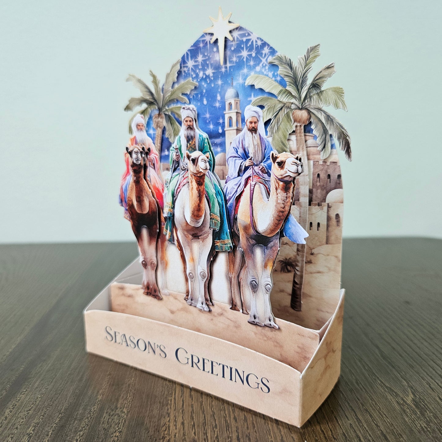 A 3D pop-up Christmas card on a wooden table against a light gray wall. The card features an illustration of three wise men on camels under a starry night sky, with palm trees and a stable in the background.