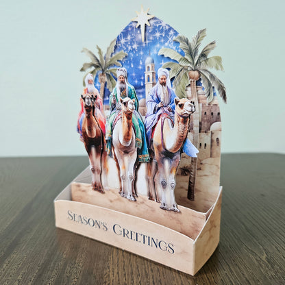 A 3D pop-up Christmas card on a wooden table against a light gray wall. The card features an illustration of three wise men on camels under a starry night sky, with palm trees and a stable in the background.