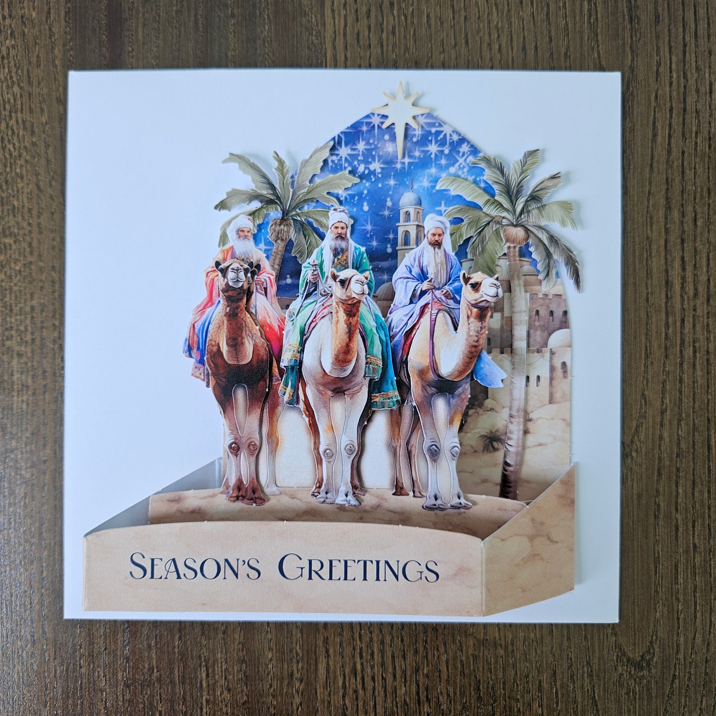 A square greeting card with a 3D pop-up design showing three camels carrying a star, set against a blue night sky with palm trees and a desert city. The right side of the card folds out to read 'SEASON'S GREETINGS' in vertical text, placed on a brown wooden surface.