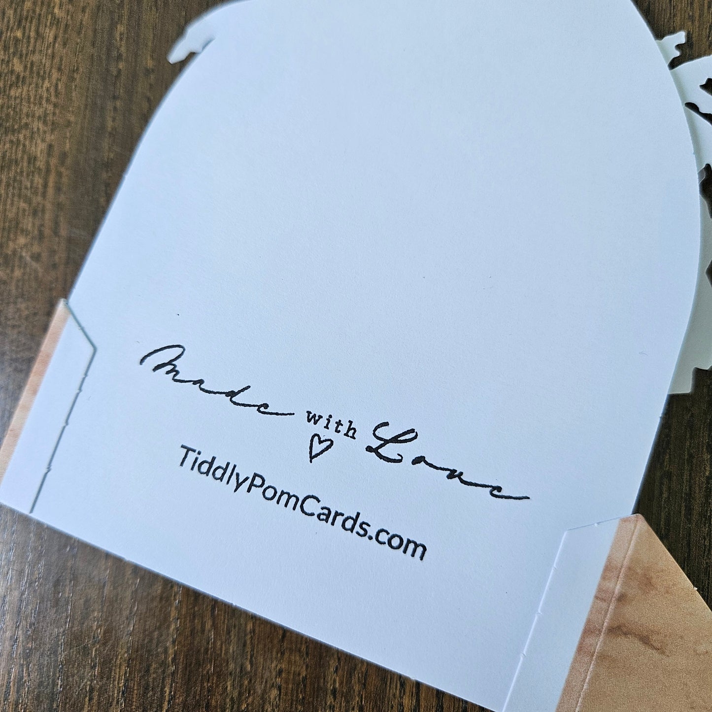 A white pom pom card with a scalloped edge, featuring the text 'Made with Love' and 'TiddlyPomCards.com' printed in black, resting on a wooden surface.