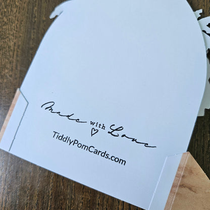 A white pom pom card with a scalloped edge, featuring the text 'Made with Love' and 'TiddlyPomCards.com' printed in black, resting on a wooden surface.