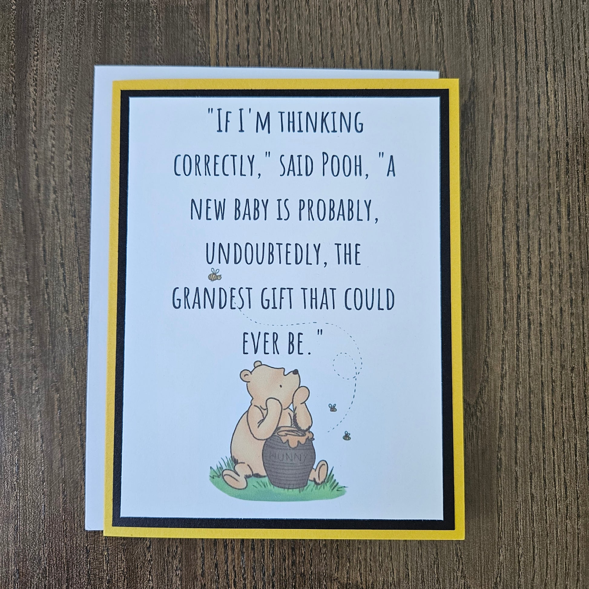 A square greeting card featuring a quote about Pooh and a honey bear illustration, photographed on a wooden surface. The card has a white center, a yellow border, a black inner border, and a white backing card.