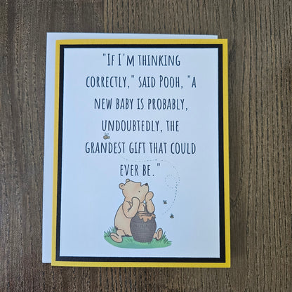 A square greeting card featuring a quote about Pooh and a honey bear illustration, photographed on a wooden surface. The card has a white center, a yellow border, a black inner border, and a white backing card.