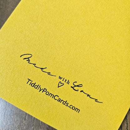 A close-up of a bright yellow card with black printed text reading 'Made with Love' and 'TiddlyPomCards.com' on a dark wood surface.