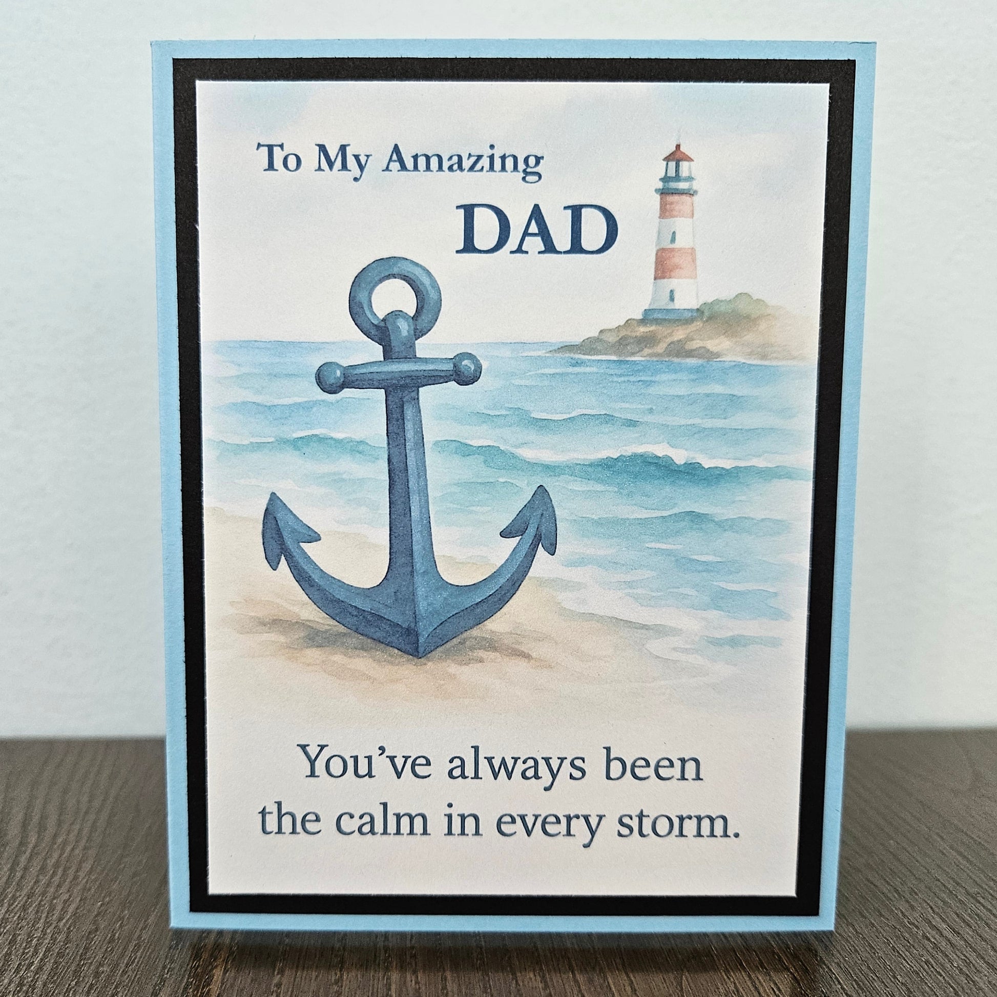 A2 greeting card with a light blue border stands on a dark wood surface against a light gray wall. The front shows a watercolor-style anchor over ocean waves, with a lighthouse in the distance and the text, 'To My Amazing Dad' and 'You've always been the calm in every storm.
