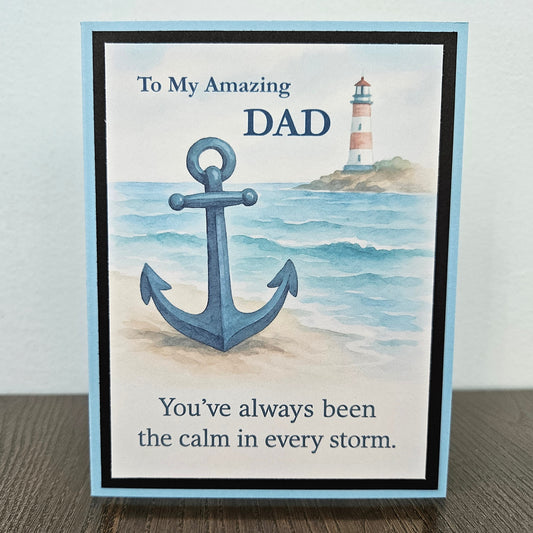 A2 greeting card with a light blue border stands on a dark wood surface against a light gray wall. The front shows a watercolor-style anchor over ocean waves, with a lighthouse in the distance and the text, 'To My Amazing Dad' and 'You've always been the calm in every storm.