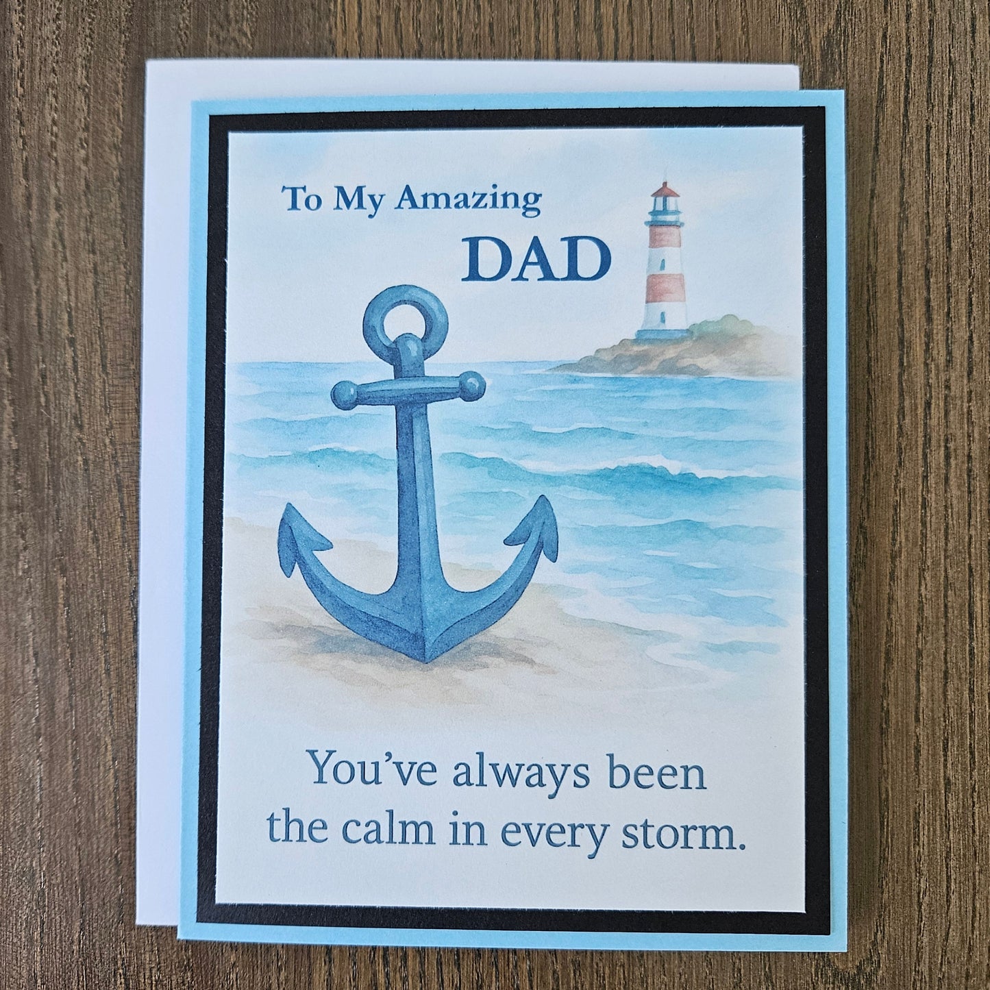 A2 greeting card for Dad featuring a nautical design with a large blue anchor, a lighthouse, and the text 'To My Amazing DAD' and 'You've always been the calm in every storm.' The card is photographed on a brown wood surface.