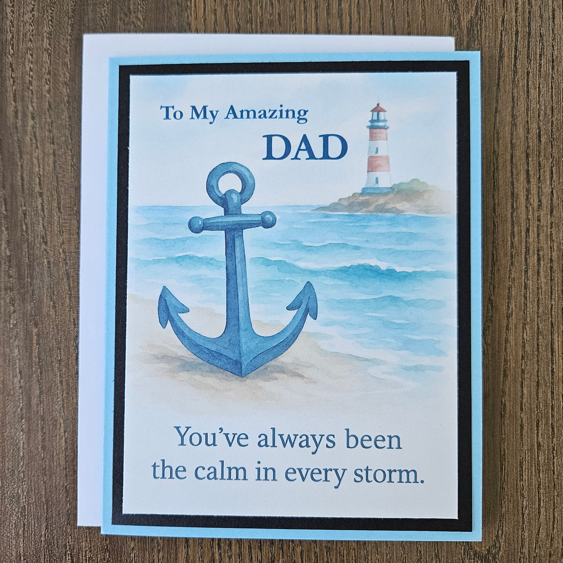 A2 greeting card for Dad featuring a nautical design with a large blue anchor, a lighthouse, and the text 'To My Amazing DAD' and 'You've always been the calm in every storm.' The card is photographed on a brown wood surface.