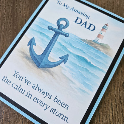 A close-up of a printed card or gift tag with a nautical design, featuring a large blue anchor over a watercolor-style ocean scene with a lighthouse. Text on the card reads, 'To My Amazing DAD' and 'You've always been the calm in every storm.