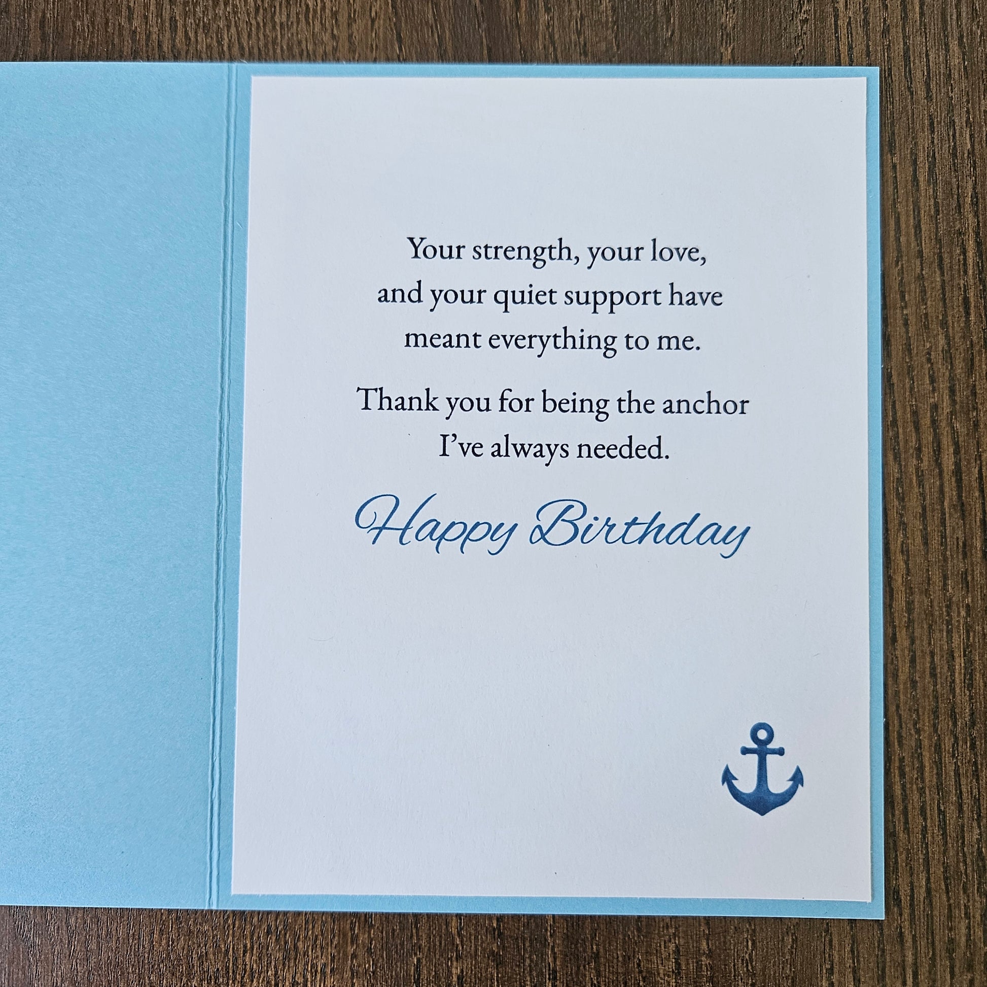 A square birthday card with a light blue envelope, featuring a white front insert with a navy blue anchor icon and the text 'Happy Birthday' and a message of support, photographed on a wooden surface.