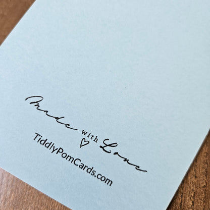 A close-up of a white card printed with the words 'Made with Love', a small heart, and 'TiddlyPomCards.com' in black script, placed on a wooden surface.