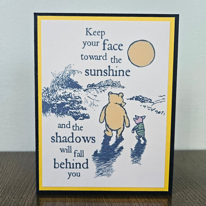A square handmade greeting card featuring a black and navy blue illustration of Winnie the Pooh and Piglet walking toward a yellow sun, with the text 'Keep your face toward the sunshine and the shadows will fall behind you.' The card has a white center, a yellow border, and a black outer frame, standing on a dark wooden surface against a light gray wall.