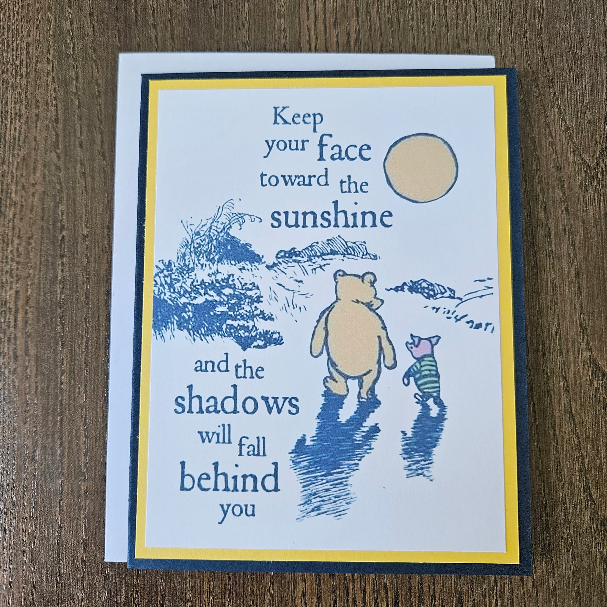 A square greeting card with a blue and yellow border, featuring a blue line drawing of Winnie the Pooh and Piglet walking outdoors, with the quote 'Keep your face toward the sunshine and the shadows will fall behind you,' shown on a wooden surface.