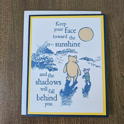 A square greeting card with a blue and yellow border, featuring a blue line drawing of Winnie the Pooh and Piglet walking outdoors, with the quote 'Keep your face toward the sunshine and the shadows will fall behind you,' shown on a wooden surface.