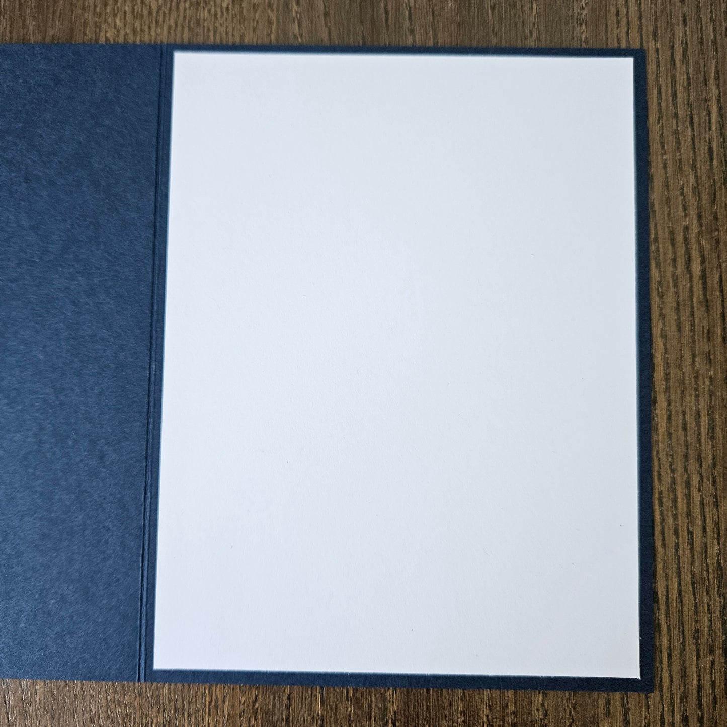 A square white card or paper insert with a dark navy border, placed on top of a textured navy fabric cover on a brown wood-grain surface.