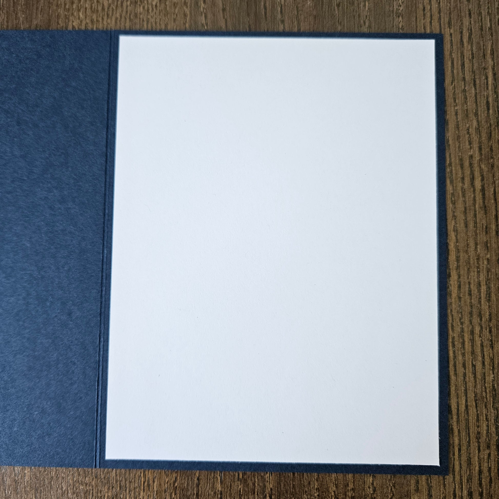A square white card or paper insert with a dark navy border, placed on top of a textured navy fabric cover on a brown wood-grain surface.