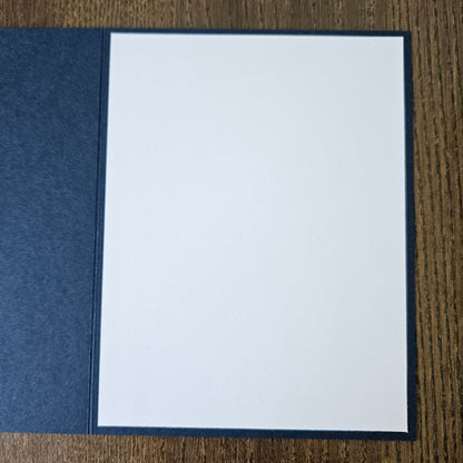 A square white card or paper insert with a dark navy border, placed on top of a textured navy fabric cover on a brown wood-grain surface.