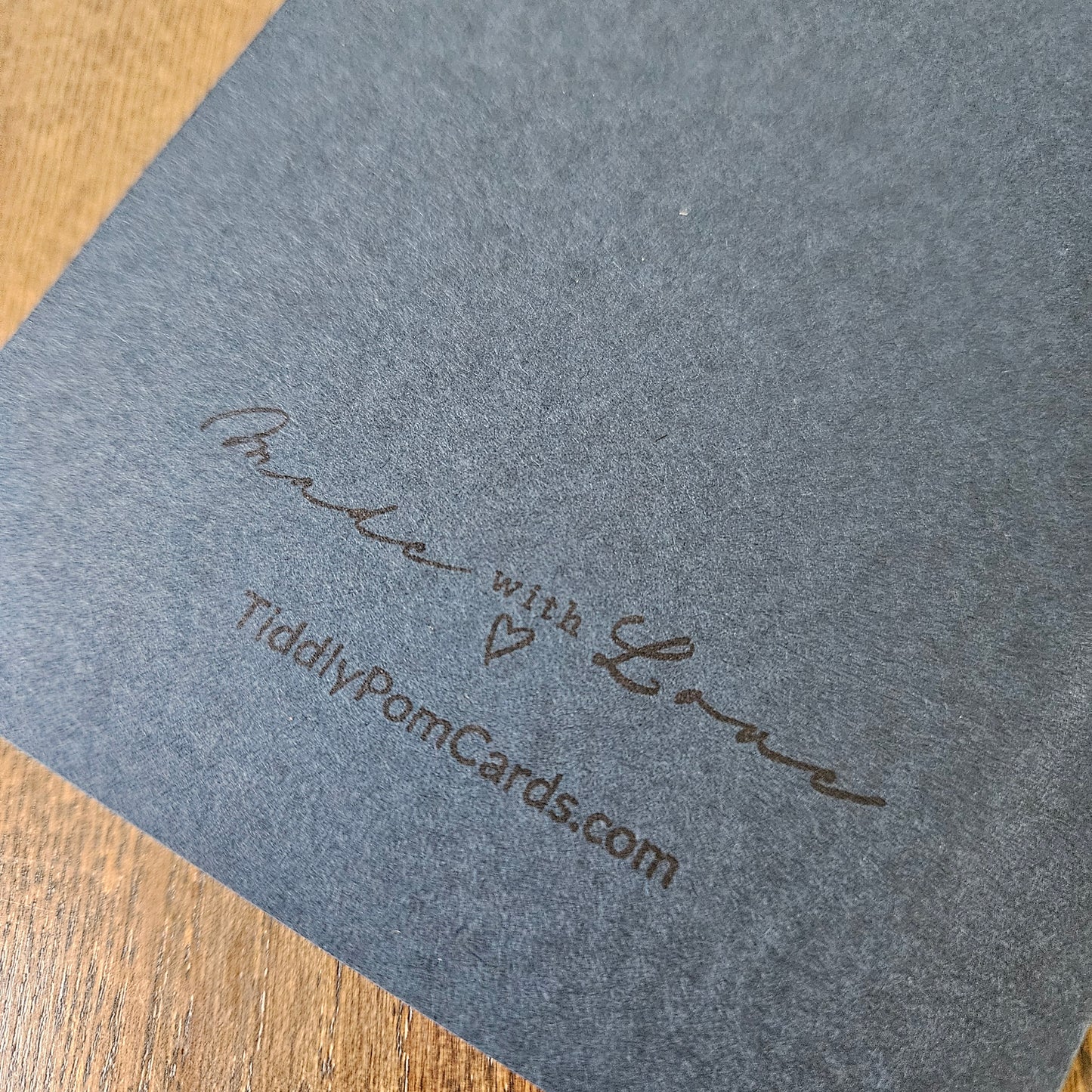 A close-up of a dark gray textured card or paper sheet with embossed script reading 'With Love' and 'TiddlyPomCards.com', placed on a wooden surface.