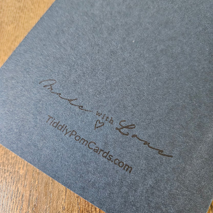 A close-up of a dark gray textured card or paper sheet with embossed script reading 'With Love' and 'TiddlyPomCards.com', placed on a wooden surface.